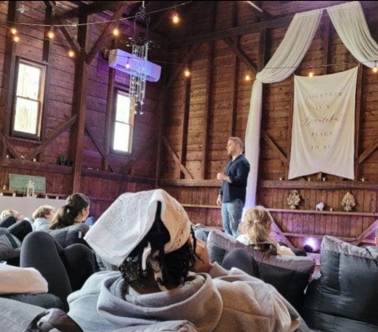 Mike James speaking to a seated audience in a barn event space with string lights and a fabric banner.
